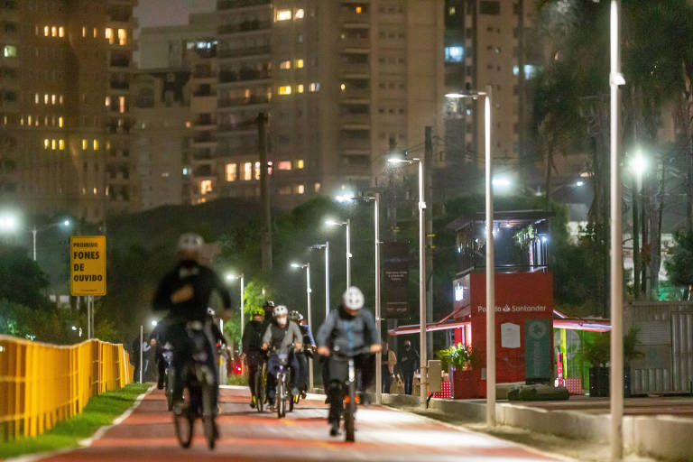 Ciclovia com vários ciclistas se exercitando à noite. Há vários postes de iluminação ao longo da ciclovia. Ao fundo, edifícios residenciais com janelas iluminadas.
