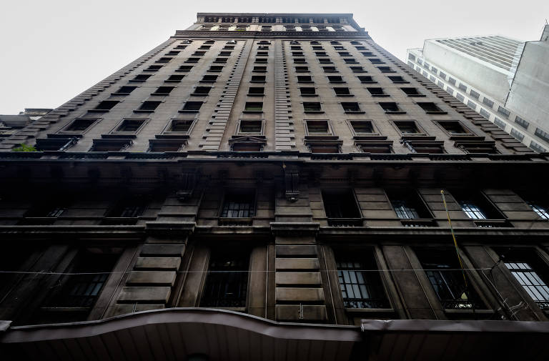 A imagem mostra a fachada de um edifício alto， vista de baixo para cima. O prédio tem uma arquitetura clássica， com detalhes em relevo e janelas emolduradas. O céu está nublado， e outros edifícios menores podem ser vistos ao fundo.
