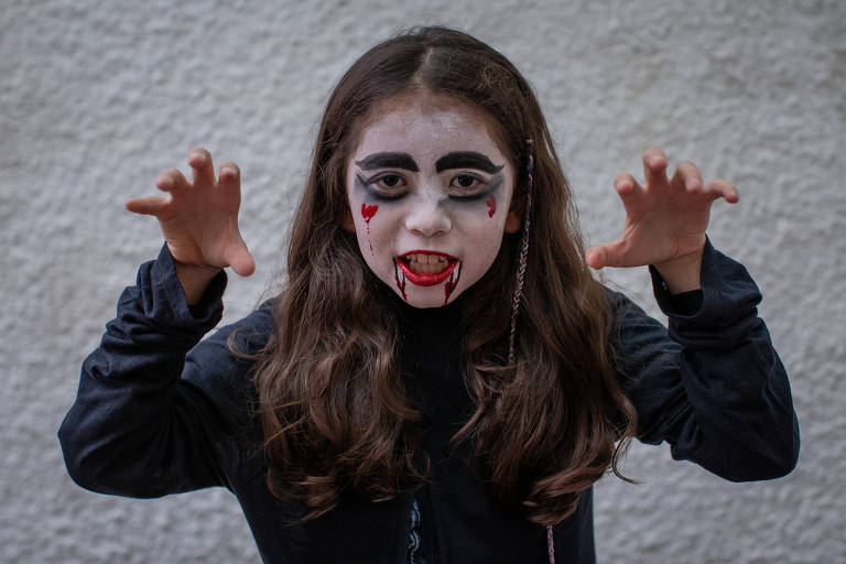 A imagem colorida mostra uma criança, de cabelos compridos castanhos, com o rosto pintado de vampiro, fazendo gestos. Seu rosto está coberto de pancake branco, com detalhes desenhados com pancake preto.