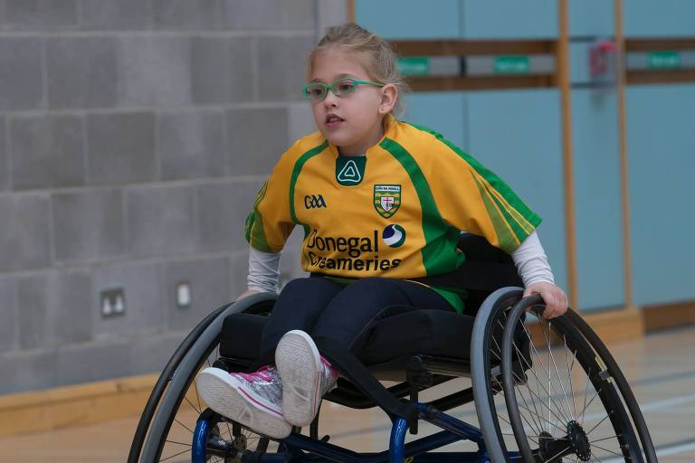 Foto mostra criança com camisa amarela e óculos em cadeira de rodas dentro de quadra esportiva, usando as mãos para mover as rodas da cadeira