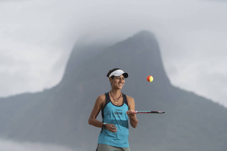 Joana Cortez, com blusa regata azul e viseira branca, bate na bola com a raquete para cima; atrás dela tem um morro envolto por neblina