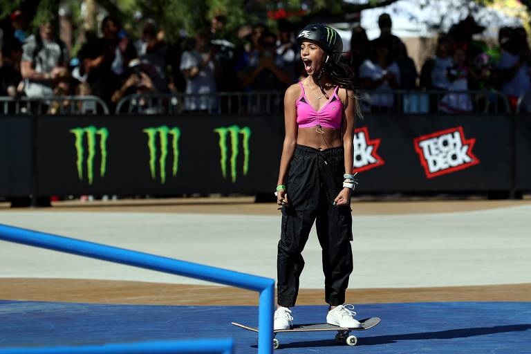 Rayssa, com capacete, top rosa e calça escura, comemora gritando sobre o skate