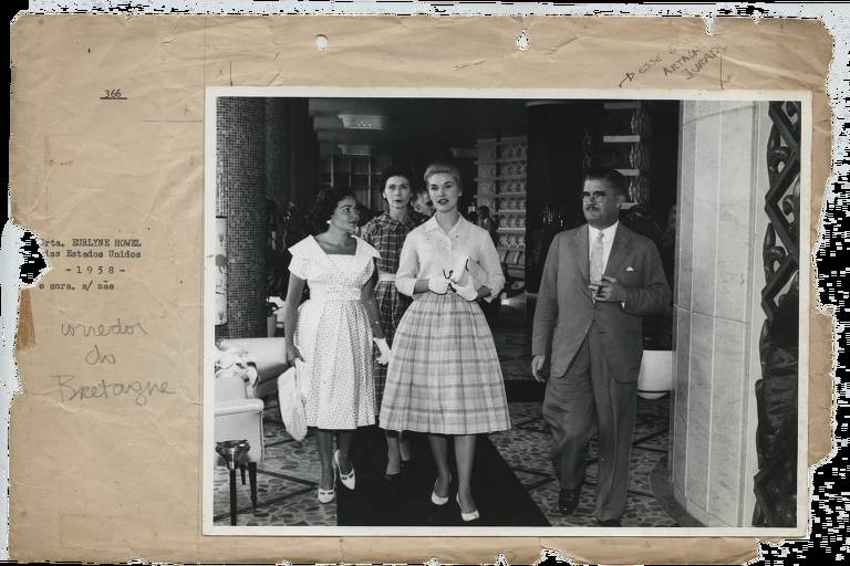 foto em preto e branco mostra três mulheres usando roupas com saias rodadas; a que vai à frente é a miss, uma mulher loura com saia xadrez, camisa de botão, usando luvas e segurando uma bolsa sob o braço esquerdo; ao seu lado, fora da passadeira por onde elas circulam, um homem de óculos e bigode, vestindo terno