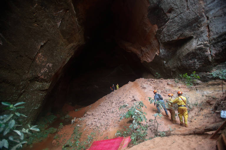 Três homens estão de pé, vestindo macacão e capacete, escalam a entrada da gruta. Ao fundo, já dentro da gruta, vê-se mais três homens, também uniformizados