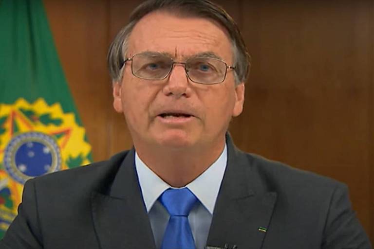 Bolsonaro, de óculos, sentado, com bandeiras ao fundo