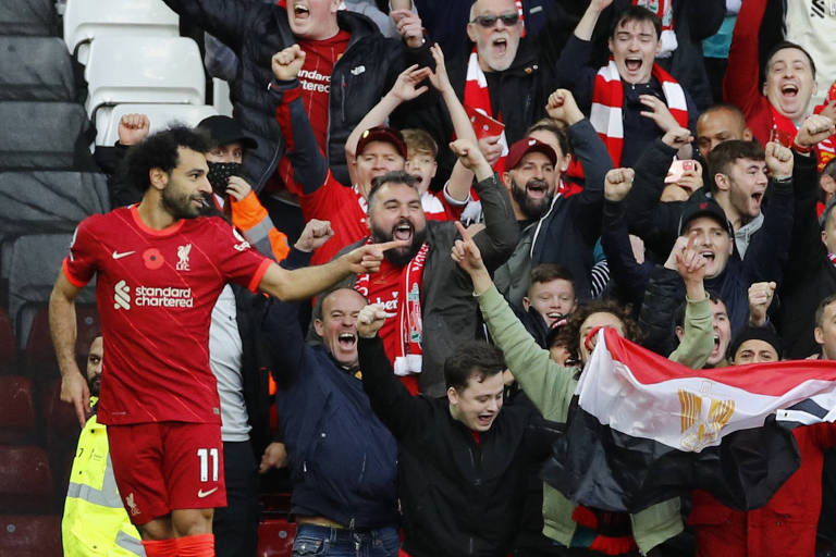 Mohamed Salah está à esquerda da foto, de frente, apontando o indicador esquerdo para o lado direito da foto. Ao fundo, pessoas na arquibancada