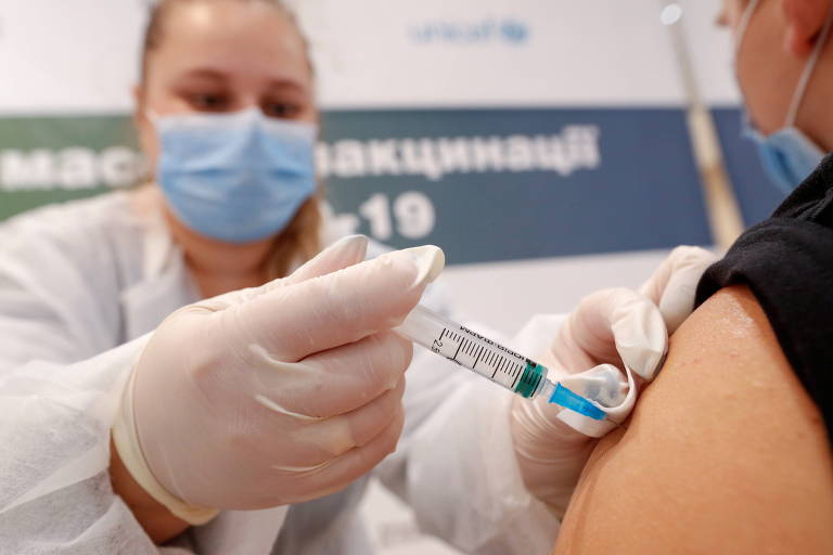 Agente de saúde de luva e máscara aplica injeção no braço esquerdo de pessoa branca com a manga da camiseta preta arregaçada