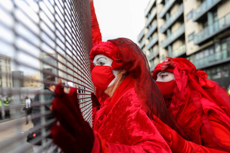 Duas moças todas cobertas de vermelho vivo, com máscaas vermelhas, olham por entre varas de uma cerca de metal
