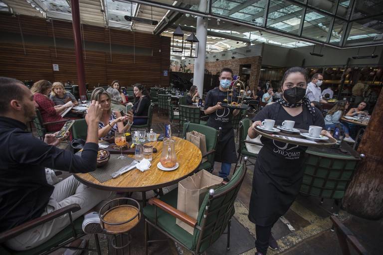 Uma jovem com roupas pretas está andando em um restaurante com uma bandeja de café nas mãos, clientes do local tiram fotos dela