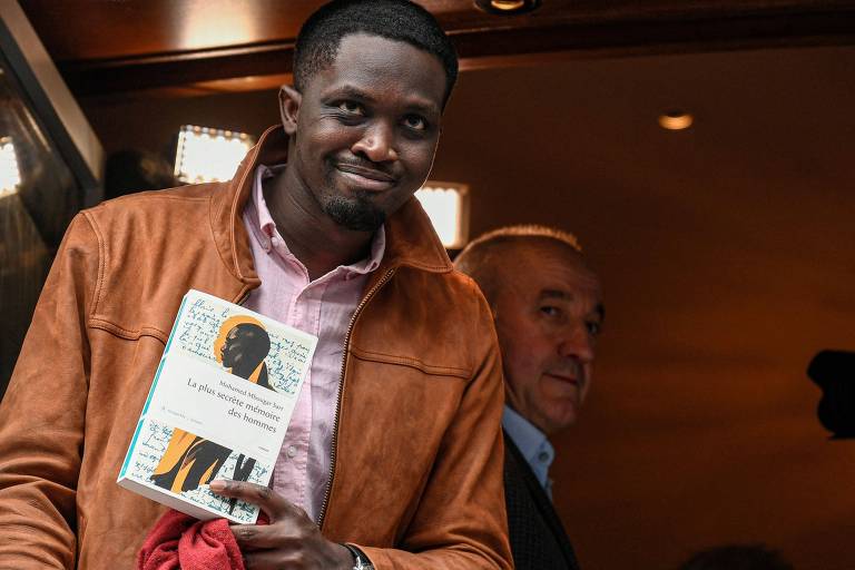 Homem negro usa um casaco de couro marrom, sorri e segura um livro em suas mãos