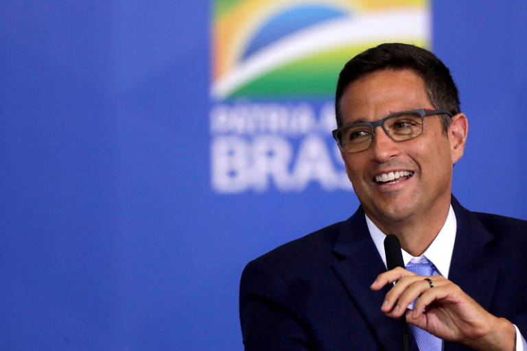 Homem de terno escuro, cabelo escuro curto e óculos retangular sorri enquanto segura microfone em frente a fundo azul com bandeira do brasil