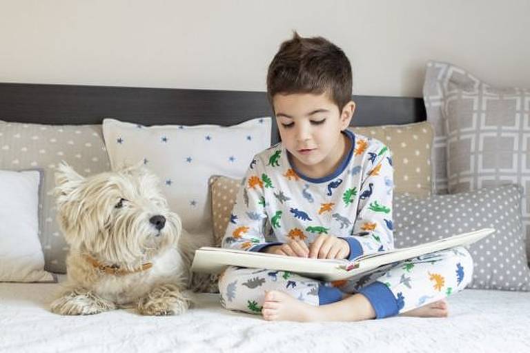 uma criança sentada em uma cama lendo um livro. Ao lado, um cachorro.