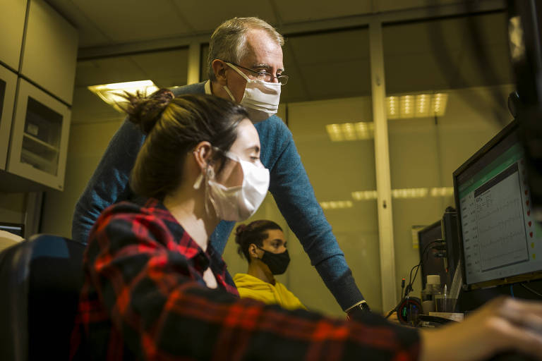 Foto mostra uma mulher, sentada, de camisa xadrez vermelha e usando máscara, mexendo em um computador. Ao lado dela, um homem de camisa azul e máscara, olha para a tela do computador, em pé. Ao fundo, outro homem parece mexendo em outro computador.