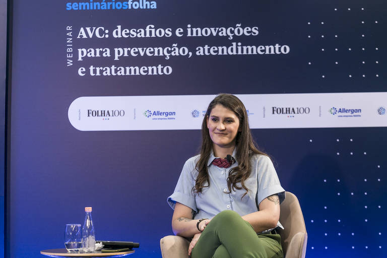Ana Bottallo aparece sentada em uma poltrona. Ao lado dela, uma mesa com uma garrafa d'água. Atrás, um painel tem os dizeres "AVC: desafios e inovações para prevenção, atendimento e tratamento.