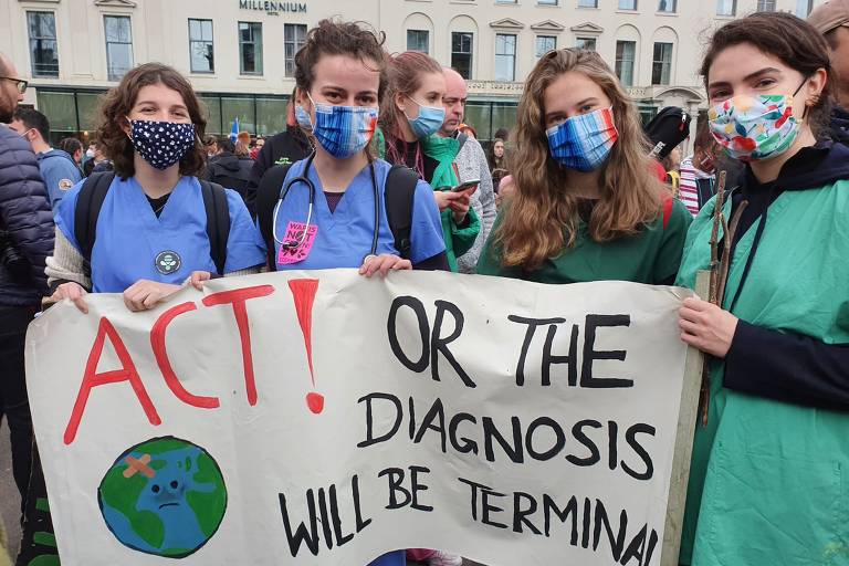 Quatro moças com uniformes de médicas e máscaras seguram cartaz dizendo Aja ou o diagnóstico será terminal