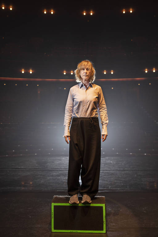 Uma mulher branca e loira está em pé em cima de um cubo no palco escuro de um teatro