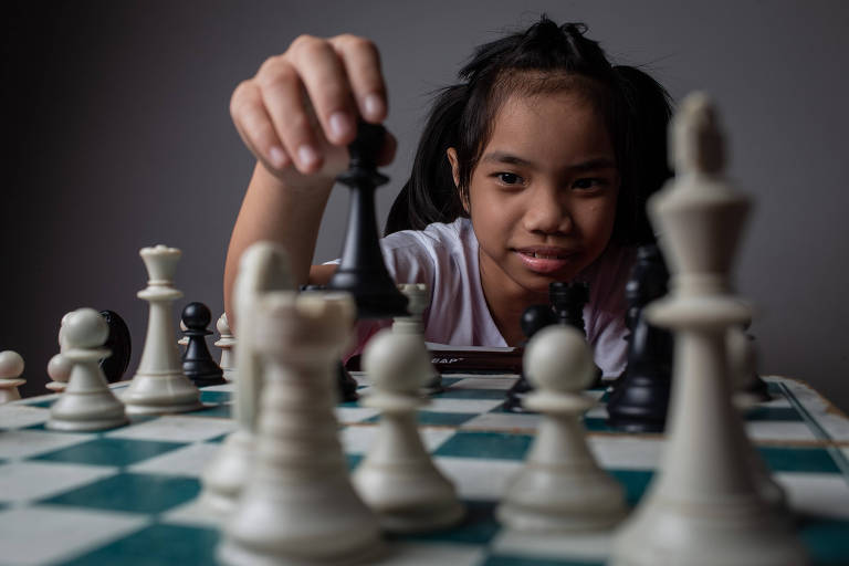 A garota Elisa Feng, 8, segura uma peça de xadrez e aparece em segundo plano, atrás de um tabuleiro com o jogo em andamento