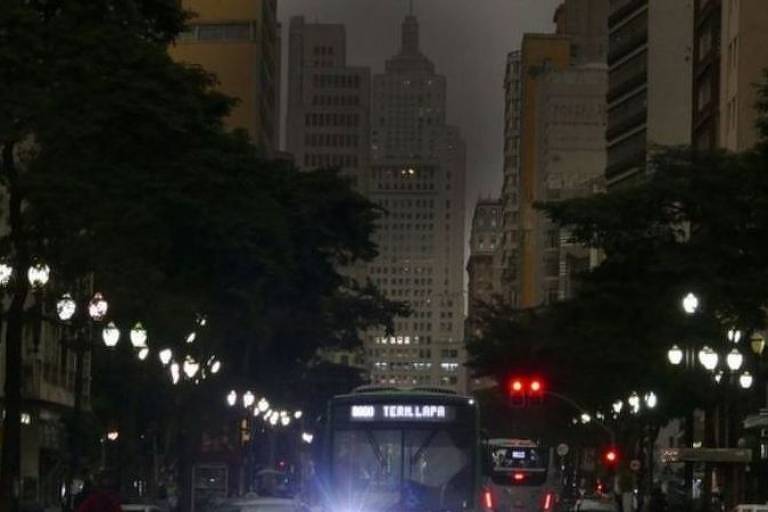 Ônibus no centro da foto e rua iluminada por postes ao redor; ao fundo e aos lados, grandes prédios