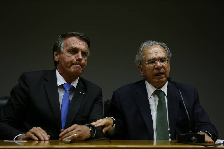 O presidente Jair Bolsonaro (esq.) e o ministro Paulo Guedes, sentados durante entrevista, com o ministro segurando o braço do presidente 