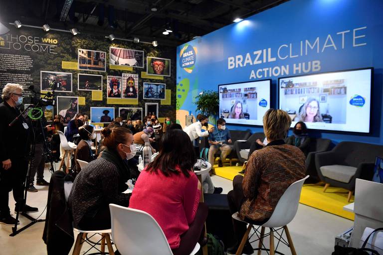 Pessoas sentadas em sala de conferência; no canto direito, duas telas; do lado direito, mais acima, é possível ler o nome da sala: Brazil Climate Action Hub