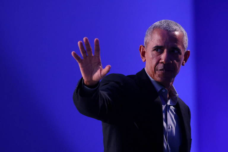 De paletó escuro em frente a um fundo azul escuro, com parte do rosto iluminada, Obama estende a mão esquerda para a frente
