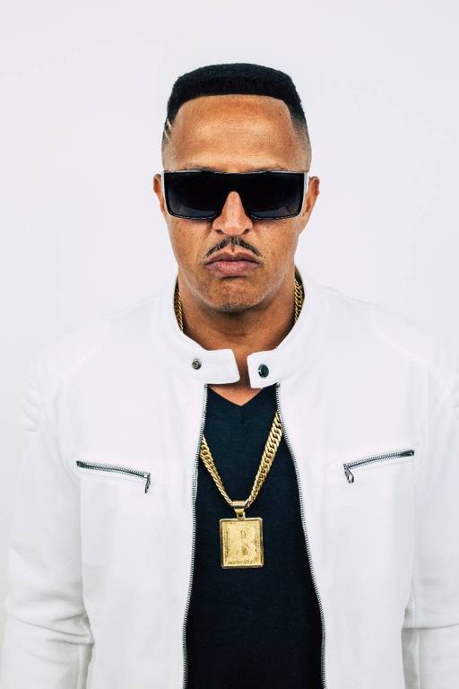 Um homem negro em pé, de óculos escuros, camiseta preta, jaqueta branca e corrente de ouro no pescoço, posa com a cara fechada, sem sorrir