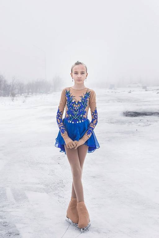  Menina usa vestido azul, típico de patinadoras, e patins. Ela está em um ambiente aberto, onde só se observa neve, gelo e algumas árvores ao fundo.