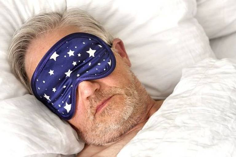 Imagem em close mostra homem crisalho com venda nos olhos deitado em uma cama
