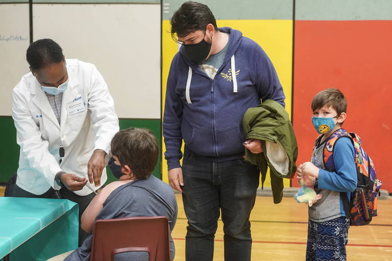Agente de saúde em pé mostra vacina para criança sentada de costas, com camiseta com a manga arregaçada para receber a dose; ao lado, o pai, em pé, e uma criança, também em pé