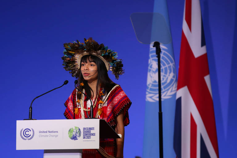 Em frente a um fundo azul, jovem indígena Txai Suruí discursa usando um cocar e roupas típicas na cor vermelha. No púlpito branco, estão a logo da COP26 e da UNFCCC.