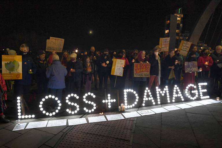 À noite, pessoas seguram cartazes atrás de sinal iluminado com as palavras "perdas e danos"