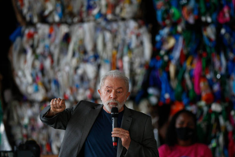 Lula está de paletó e camiseta escuros e fala ao microfone. Ao fundo, imagem colorida (lixo reciclado, mas está desfocado)