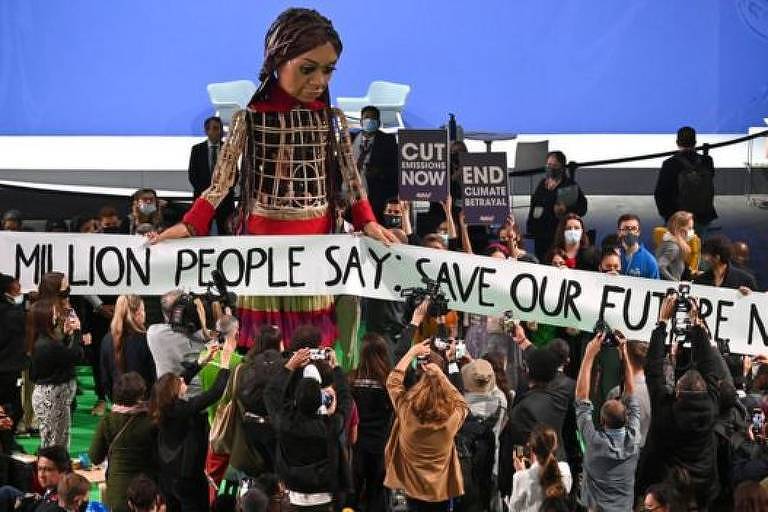 Em destaque, há uma boneca gigante e um cartaz escrito "1,8 million people say save our future now". Atrás dela, há um grupo de manifestantes e, à frente, um grupo de pessoas filmando e fotografando