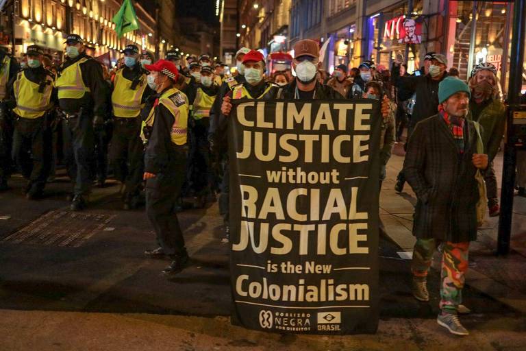 Em destaque, um homem segura um cartaz escrito: climate justice without racial justice is the news colonialism. Ao fundo, se vê um grupo de manifestantes.