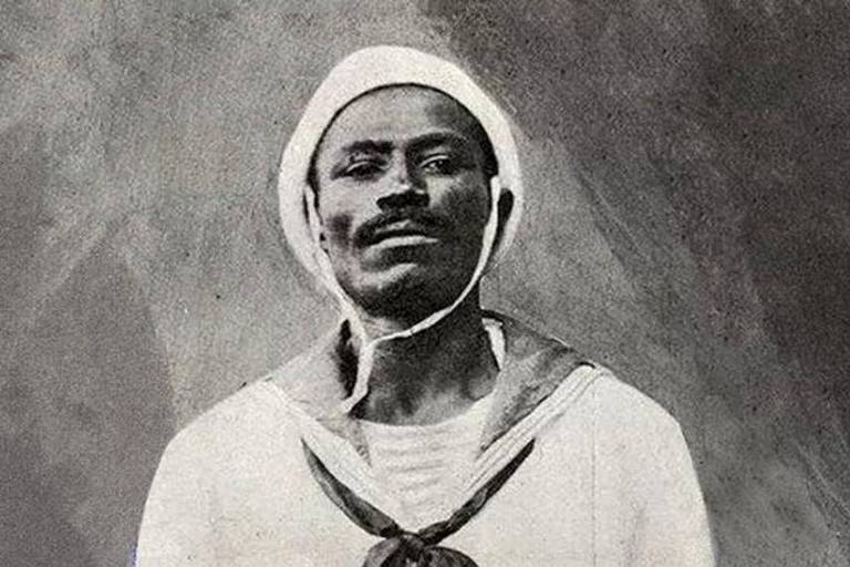 Homem negro, de aparência séria, bigode preto e uniforme branco de marinheiro, posa para foto. A foto é antiga, em preto e branco