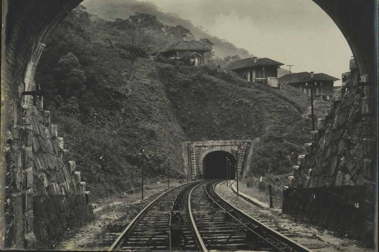 Imagem em preto e branco mostra entrada de um túnel e trecho de uma estrada de ferro