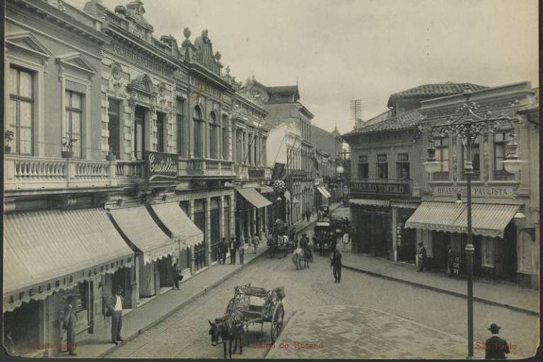 Imagem em preto e branco mostra mostra antigo Largo do Rosário. Em uma rua, se vê construções antigas e transporte feito por tração animal