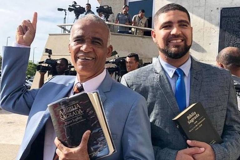 Dois homens usando terno e gravata, cada um deles segurando uma Bíblia na mão. A esquerda esta o mais velho, grisalho e a direita esta o filho, usando barba