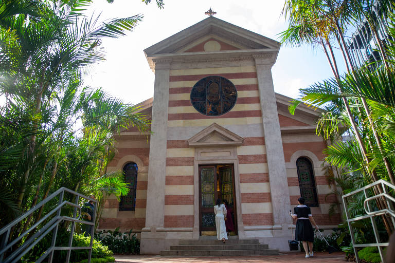 Igreja com fachada decorada com faixas lisas de cor bege alternadas com outras que imitam tijolo; ela é cercada de vegetação