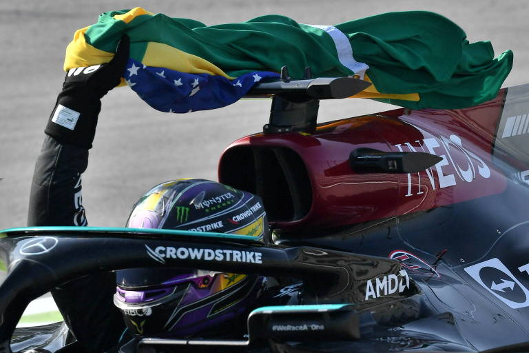 Hamilton, piloto de uniforme e capacete escuros segura no alto com a mão direita bandeira do Brasil com carro de F1 preto com detalhes em verde-água e vermelho em movimento