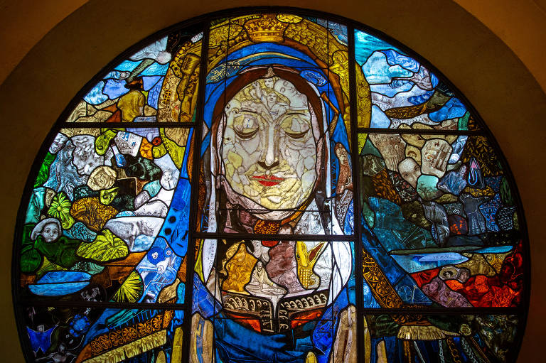 Janela circular feita em vitral colorido mostra imagem de uma santa com pequenas outras imagens ao redor