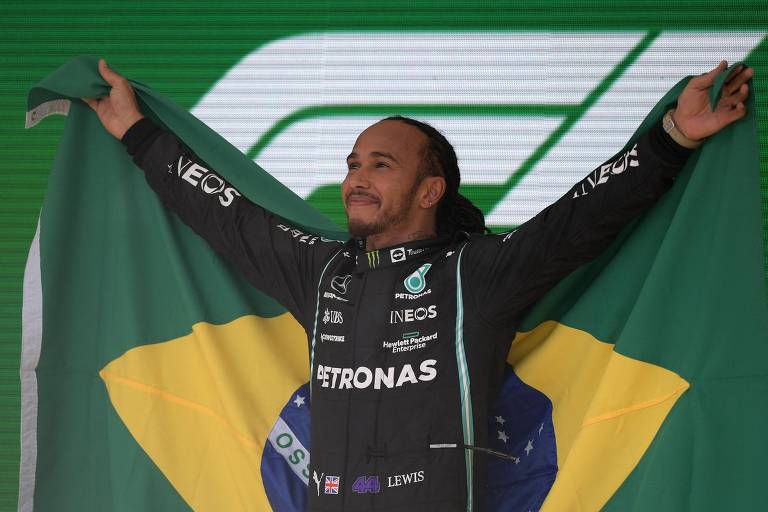 Lewis Hamilton, negro, de barba curta, estende os braços segurando por trás a bandeira do Brasil