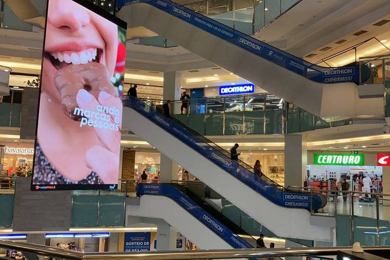 escadas rolantes em shopping, com painel digital em frente, com imagem de mulher degustando chocolate