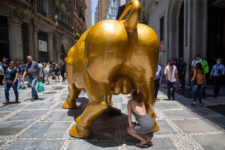 Mulher toca testículos de réplica do Touro de Wall Street, em frente ao prédio da Bolsa, no centro de São Paulo; superstição sobre escultura original associa gesto à prosperidade