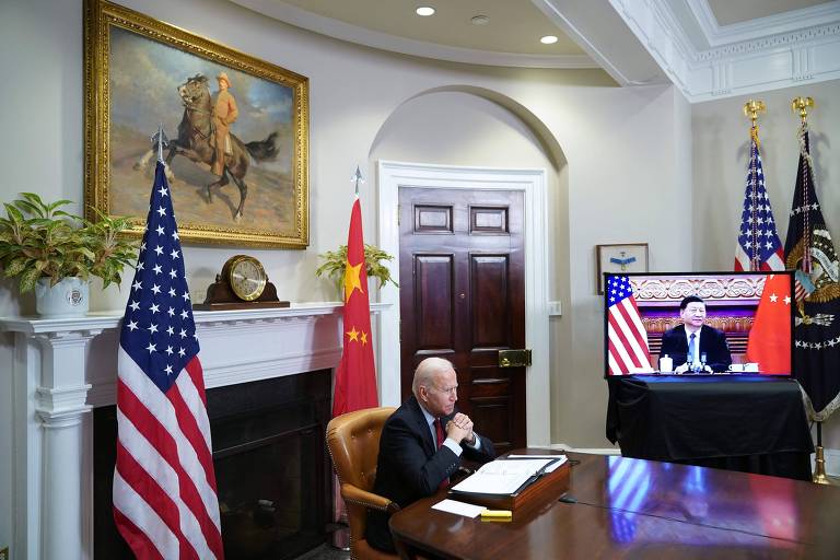 Foto mostra o presidente os Estados Unidos Joe Biden sentado em sua mesa. Ao fundos, às bandeiras de China e Japão e uma televisão com a imagem de Xi Jinping, com quem ele conversava virtualmente.