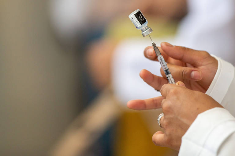 Profissional de saúde (da qual só vemos as mãos) segura uma seringa em que a agulha está enfiada em um frasco com vacina; o frasco está sendo sustentado pela agulha