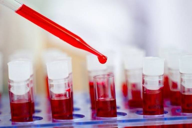Imagem em close mostra ampolas com líquido vermelho dispostas sobre uma bandeja em um laboratório. Um conta gotas despeja o líquido em um dos frascos.