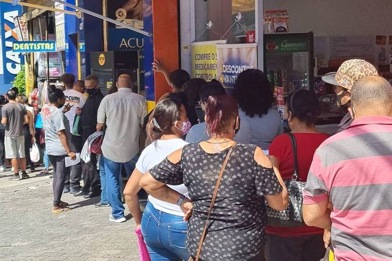 Dezenas de pessoas numa fila formada numa calçada de uma rua comercial no bairro de Itaquera, Zona Leste de São Paulo