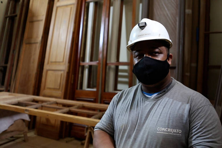em uma sala de marcenaria, com várias portas de madeira espalhadas, homem posa para foto usando máscara contra a covid-19 e capacete de proteção