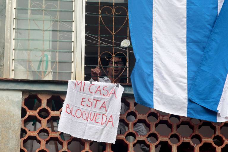 Com a casa cercada por agentes do regime cubano, Yunior García mostra, da janela, uma faixa com a frase 'minha casa está bloqueada' 